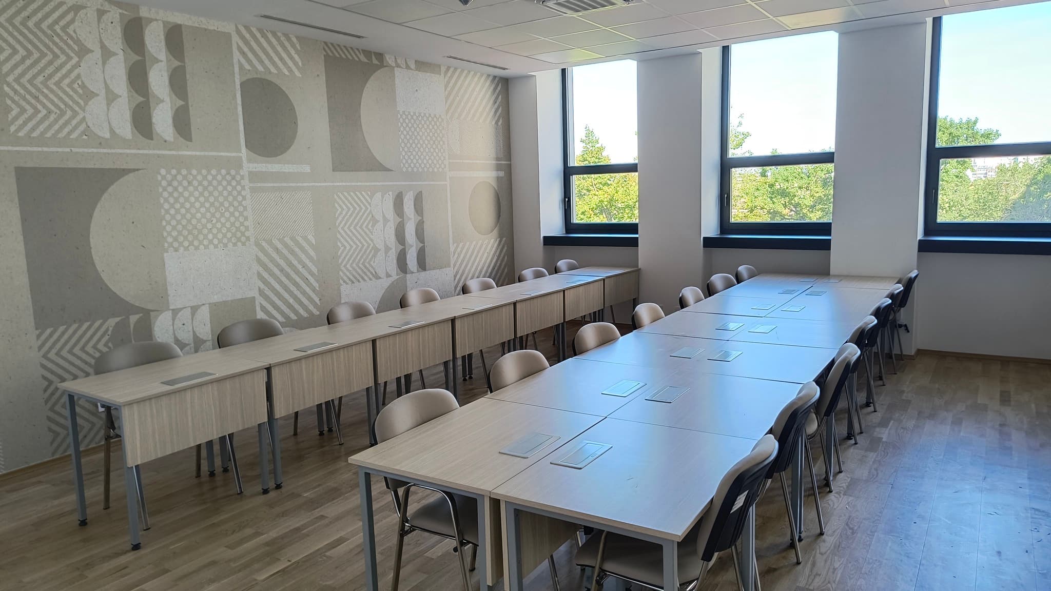 Sală de seminar la ASE București: mese din pal cu decor de stejar deschis, cu prize încastrate în blat, scaune tapițate și un perete decorat cu motive geometrice, lângă ferestre ample.