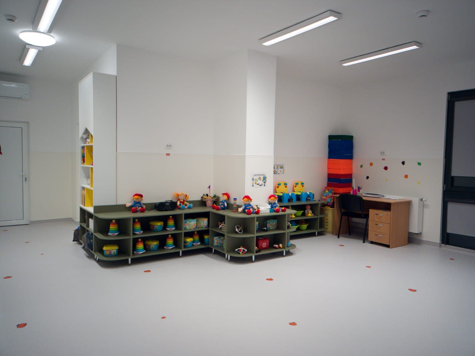 Sală de joacă de creșă cu mobilier modular jos verde, bibliotecă galbenă deschisă și birou mic pentru copii, cu jucării colorate pe rafturi.