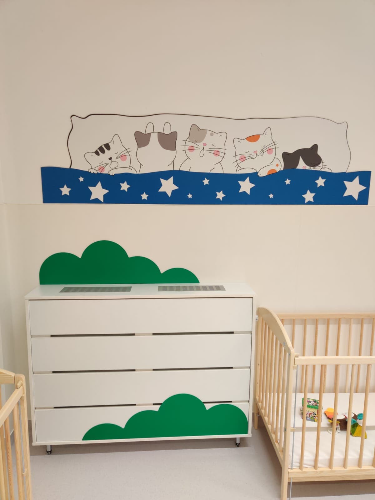 Dormitor de creșă cu pătuț din lemn natur de pin, comodă albă cu motive de norișori verzi și un mural cu pisicuțe care dorm.