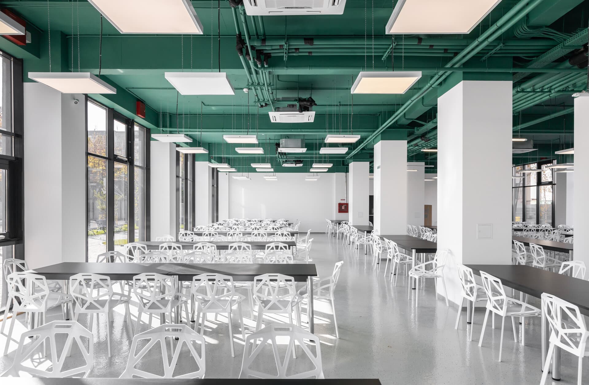 Cantina Colegiului Miron Nicolescu: tavan tehnic vopsit verde, mese negre dreptunghiulare și scaune albe geometrice din metal perforat, lângă ferestre mari.