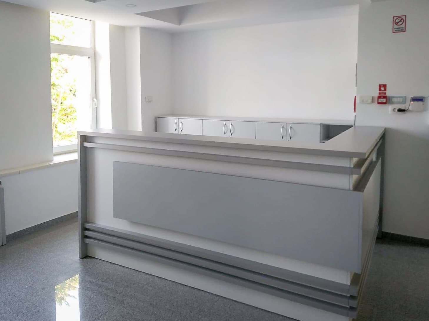 Vedere frontală a recepției dintr-un cabinet medical privat din București: birou în formă de L cu panouri gri, dulapuri joase de depozitare și pardoseală de granit lustruit.