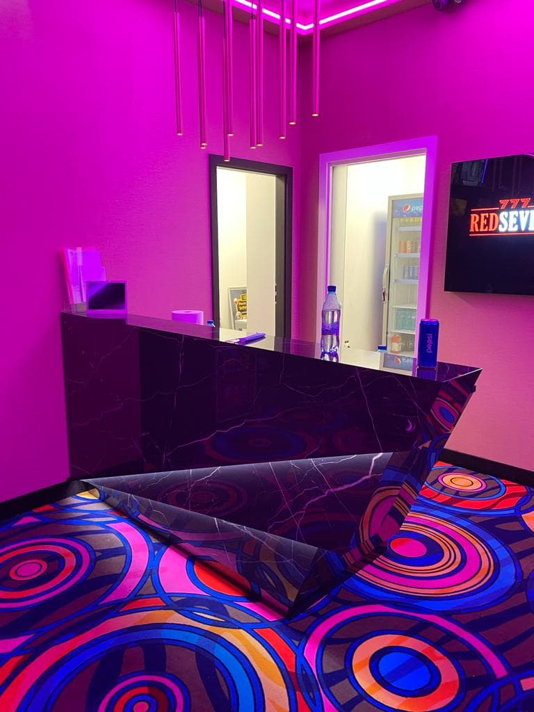Bar de recepție angular din marmură neagră lucioasă cu muchii tăiate, într-o sală de jocuri cu iluminat neon și mochetă cu model de cercuri concentrice.