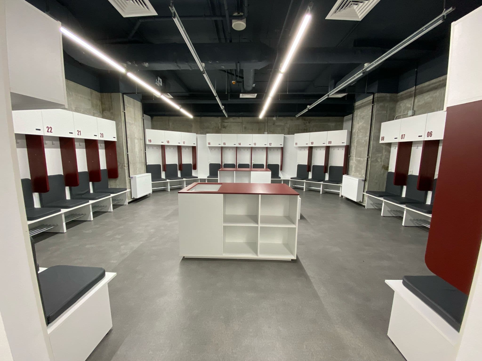 Vestiar de fotbal cu boxe albe numerotate cu accente bordo, două insule centrale de depozitare cu blat vișiniu și tavan tehnic negru cu benzi LED.