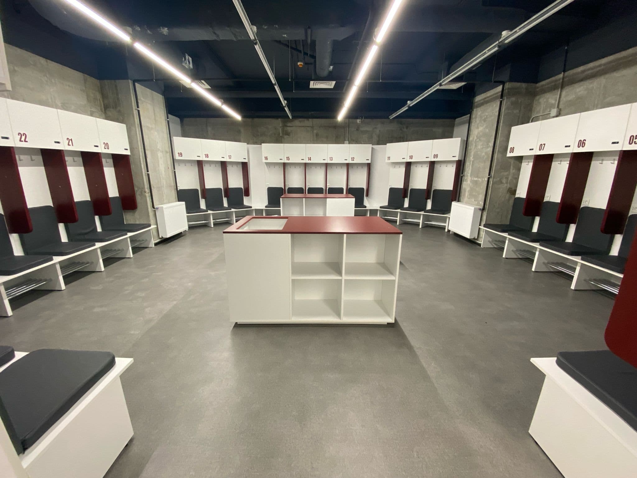 Vestiarul echipei de la Stadionul Rapid: dulapuri albe numerotate deasupra, panouri de spătar vișinii, bănci tapițate gri și un corp central de depozitare cu blat vișiniu, pe fundal de beton aparent.