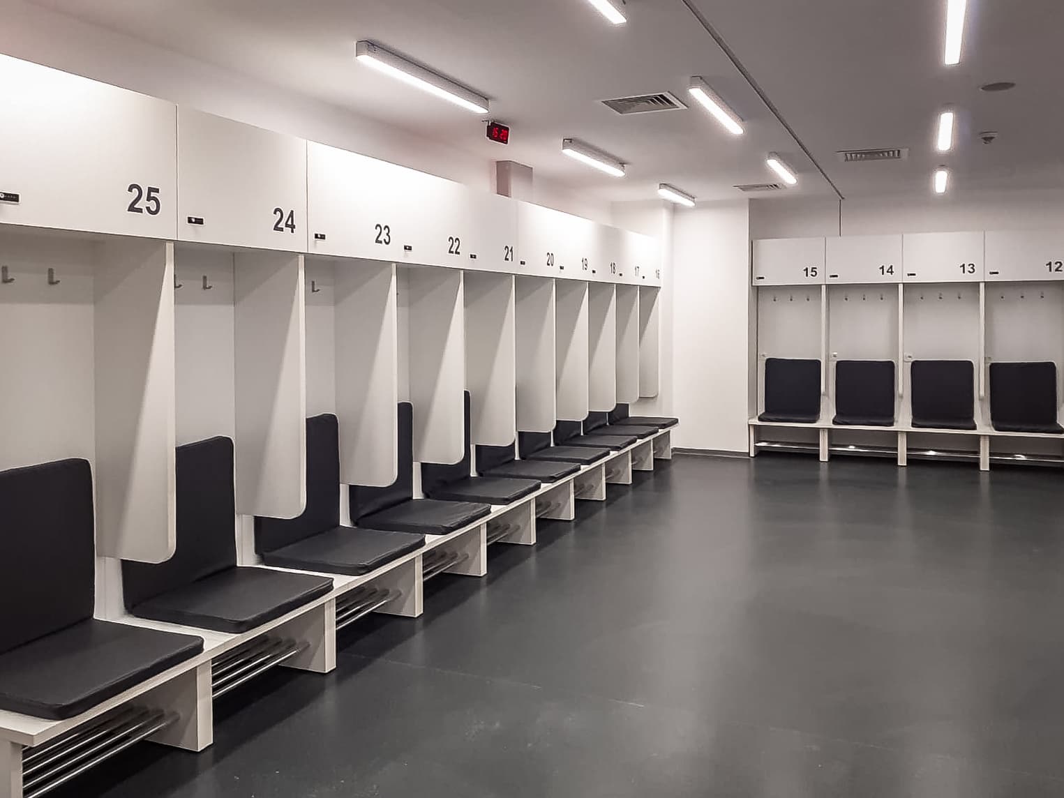 Vestiar sportiv finisat, cu boxe individuale albe numerotate, dulapuri suspendate, șezuturi tapițate negre și iluminare cu benzi LED.