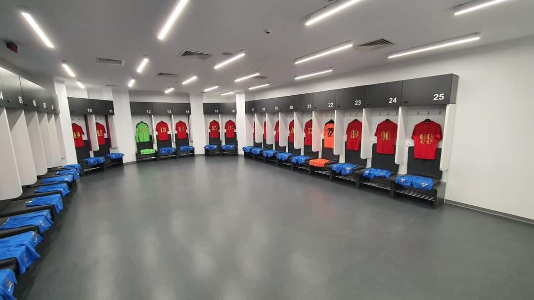 Vestiarul Stadionului Steaua pregătit de meci: tricouri roșii cu numere aurii atârnate în compartimentele numerotate, cu echipament albastru împăturit pe bănci.
